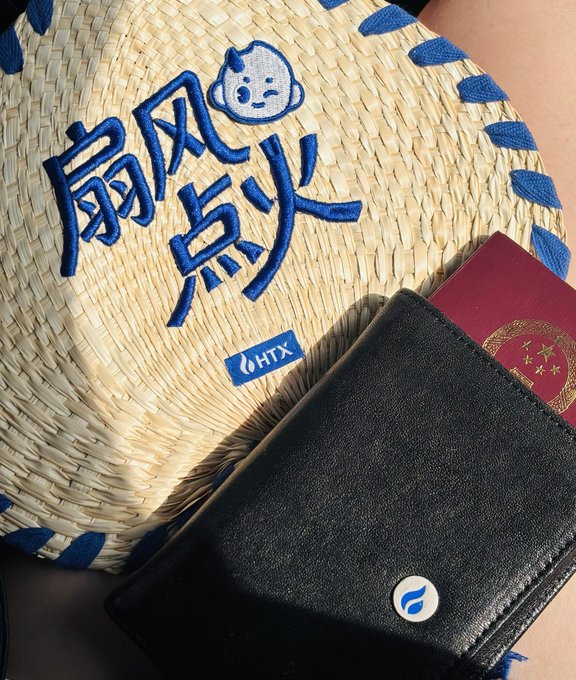 A straw hat featuring blue embroidered Chinese characters '扇风点火' (stirring up trouble) with a small baby face icon and an HTX logo, resting next to a black wallet and a Chinese passport.
