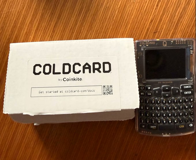 A Coinkite COLDCARD hardware wallet sits next to its white packaging box on a wooden surface, with the box displaying "COLDCARD by Coinkite" and a QR code linking to coldcard.com/docs. The image relates to the post by showing the physical self-custody Bitcoin storage device the author received two years ago to secure their investments independently.