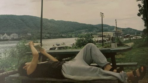 The image features a person laying on a bench, reading a book while enjoying the view of a river or lake nearby. The person appears relaxed and content as they immerse themselves in their book. In the background, there are several cars parked along the shoreline, indicating that this location is popular for outdoor activities like picnics or leisurely strolls by the water.