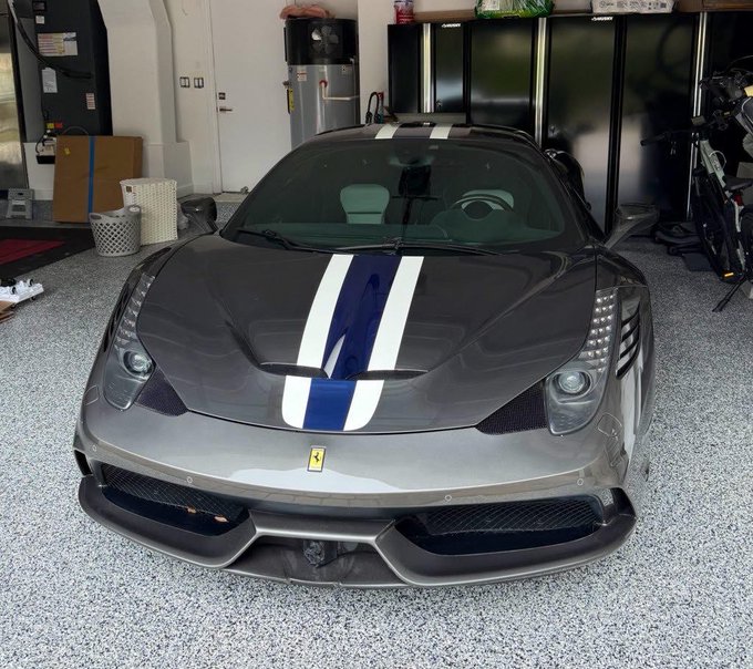 A sleek gray Ferrari with blue and white racing stripes parked in a garage, the high-performance exotic car humorously symbolizing the type of expensive splurge someone might make after cashing out Bitcoin at $58k.