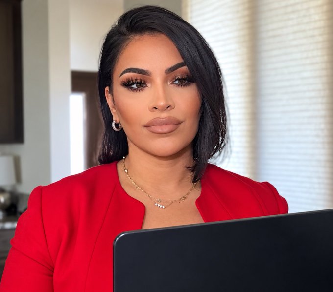 A professional portrait of a woman with dark hair wearing a red blazer and delicate gold necklace, sitting in front of a laptop.