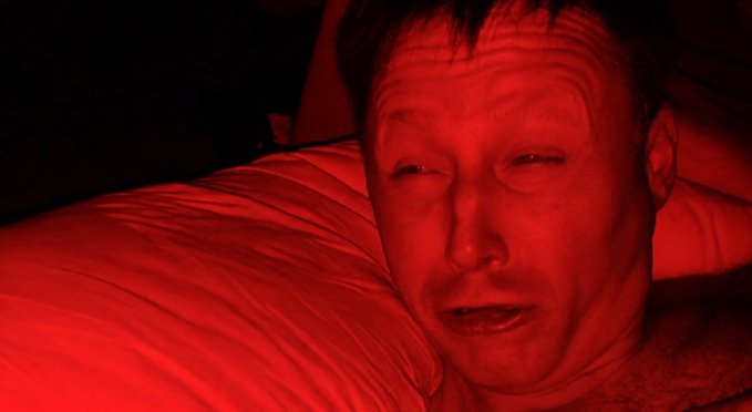 A man's face illuminated by red light as he lies on a pillow.