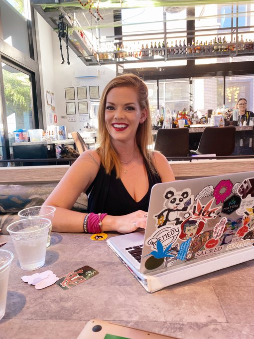The image features a woman sitting at a table with her laptop open in front of her. She has a cup of water next to her, indicating she might be working or studying for an extended period. The background shows several chairs around the dining tables, suggesting that this could be a restaurant or cafe setting. There are also other people present in the scene, possibly enjoying their time at the establishment as well.