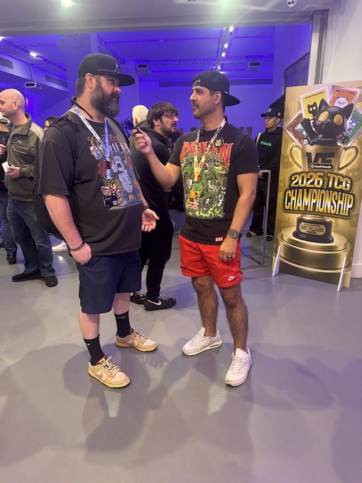 Two men in casual attire and baseball caps standing and smiling in front of a '2026 TCG Championship' banner featuring Pokemon-style artwork.