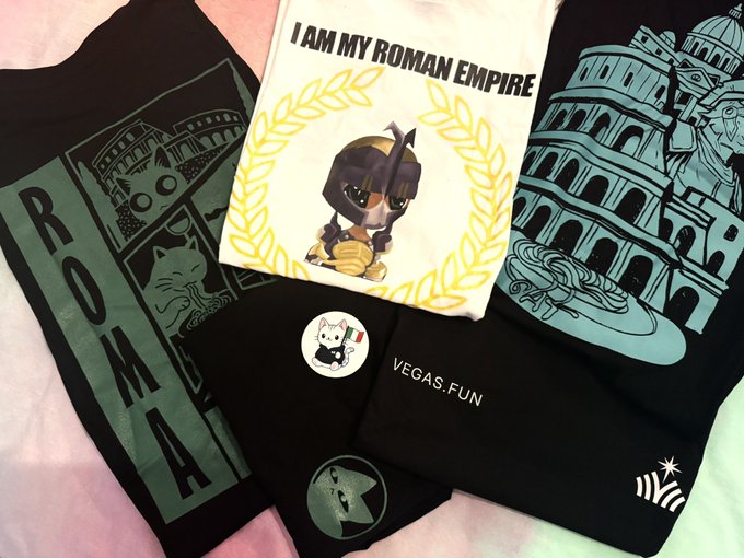 Roman-themed merchandise laid out on a pink surface, including black t-shirts with "ROMA" and Colosseum graphics, a white shirt reading "I AM MY ROMAN EMPIRE" featuring a gladiator character, and a "VEGAS.FUN" branded item.