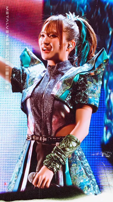 A female performer on stage wearing a teal and silver ornate costume with shoulder armor and arm gauntlets, smiling at the camera.