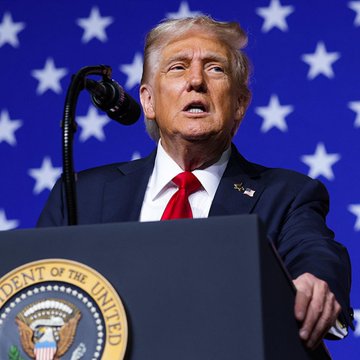 The user wants a concise 1-2 sentence caption describing the image.
I need to identify the key visual elements:
- A man (Donald Trump) speaking at a podium.
- The podium has the Presidential Seal ("PRESIDENT OF THE UNITED STATES" is partially visible, specifically "DENT OF THE UNITED").
- The background is a blue flag with white stars (US flag motif).
- He is wearing a dark suit, white shirt, and red tie.

The prompt asks for a caption describing what is in the image, but also mentions a post about the "US Strategic Bitcoin Reserve". I need to connect the image to this context if possible, or just describe the image as requested. The prompt says "Write a concise 1-2 sentence caption describing what is in this image." It also says "Include any key text...". And then it gives a post about Bitcoin. It asks "Note how the image relates to it." This implies I should
