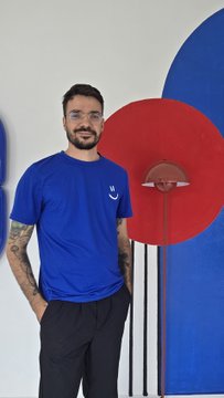 A man with short dark hair and tattoos, wearing a blue t-shirt and dark pants, stands in front of a white wall decorated with large red and blue circular shapes.