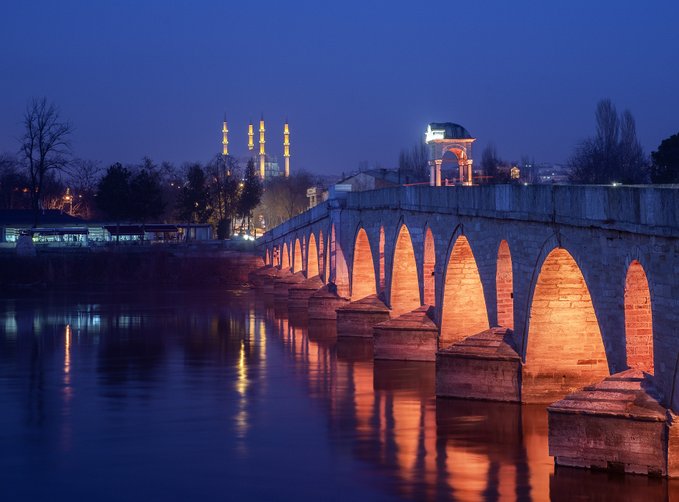 A nighttime cityscape featuring an illuminated stone arched bridge reflecting on water, with a lit mosque minarets in the background.