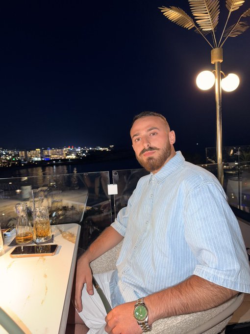 A bearded man in a light blue striped shirt poses at a rooftop bar at night, with city lights glittering across the water behind him and a stylish palm-shaped lamp overhead — setting the perfect scene for "best caption gets 3 SOL" vibes.