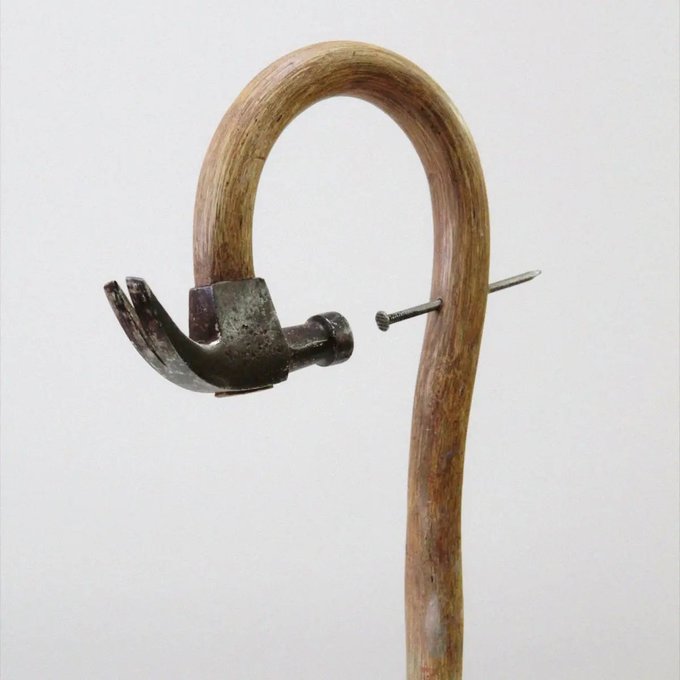 An unusual cane features a metal hammer head incorporated into its curved wooden handle, with a small metal spike visible to the side.