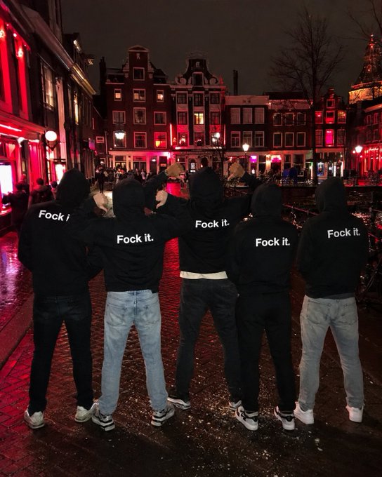 Five people in black hoodies with "Fock it." printed on the back stand together on a wet street in what appears to be Amsterdam's red light district at night, with red-lit buildings and a church dome in the background.