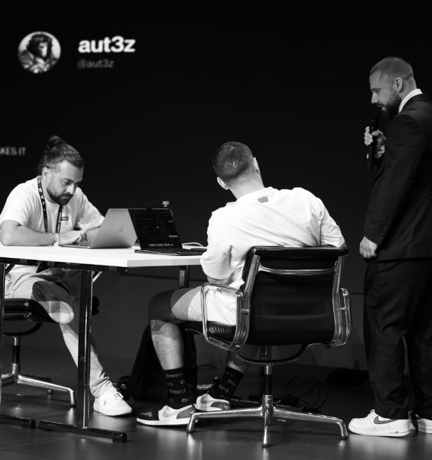 A black-and-white photo showing two people working on laptops at a table while a man in a suit holding a microphone stands to the side, with 'aut3z' branding visible on the wall.