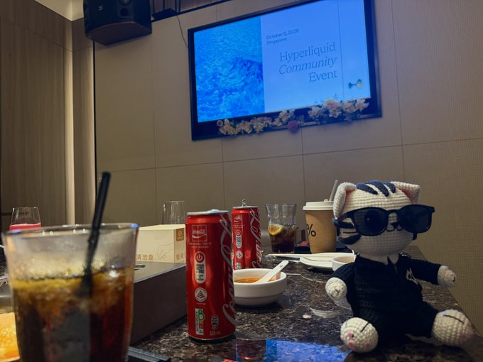 A casual gathering setup featuring Coca-Cola cans, iced drinks, and a crocheted amigurumi doll wearing sunglasses and a black jacket, with a TV displaying "Hyperliquid Community Event" dated October 5, 2025, in the background.