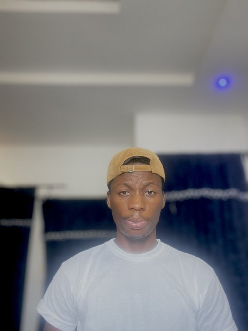 A young man wearing a white t-shirt and a tan cap worn backwards stands indoors in front of a blue curtain, looking at the camera with a slight smile.