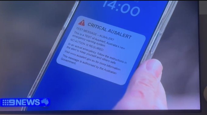A hand holds a smartphone displaying a sample "CRITICAL AUSA LERT" test message at 14:00, previewing Australia's new emergency warning system being tested on July 27 at 2pm AEST before nationwide rollout in October.
