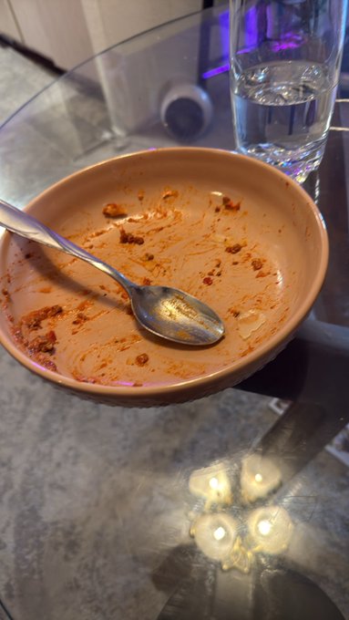 An empty, orange-stained ceramic bowl with a metal spoon sits on a glass table next to a glass of water.