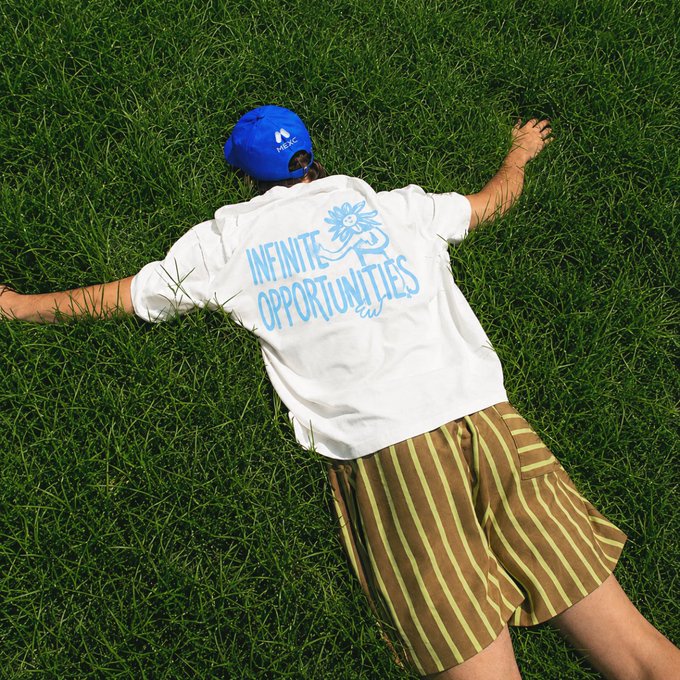 The image shows a person lying on their stomach with arms and legs spread out, wearing a white t-shirt that says "infinite opportunities" and striped shorts. The person appears relaxed and comfortable while laying down in the grassy area. The photo also includes text that reads "Therapist: Have you tried touching grass?" which suggests that the person might be experiencing stress or anxiety and is being encouraged to engage with nature as a form of relaxation, possibly by touching the grass.