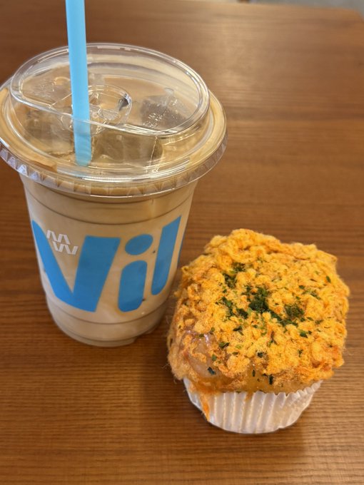 An iced coffee drink in a clear plastic cup with a blue "WAWA" logo and blue straw sits on a wooden table next to a muffin topped with orange streusel and green herbs.