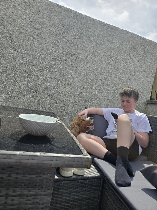 A young boy in a white t-shirt, black shorts, and black socks sitting on an outdoor wicker sofa, petting a brown dog beside a black table.