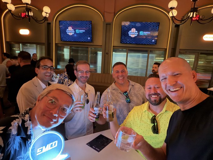 A group of six men smile and toast with drinks at an EMCD-branded event, with "EMCD" visible on a glowing sign in the foreground and on screens in the background, fitting the celebration of the EMCD and Vnish firmware party at Mining Disrupt.