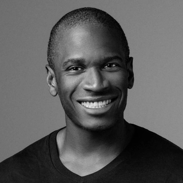 A black-and-white portrait of a smiling Black man with short hair wearing a dark v-neck shirt against a gray background.