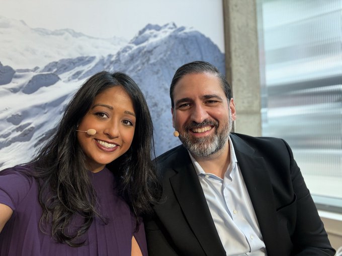 A man and woman wearing headset microphones smile at the camera, with a snowy mountain photograph hanging on the wall behind them.