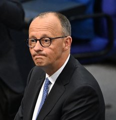 A close-up photograph of a man in a dark suit and blue tie wearing round glasses, appearing to be a coach or official at an event.