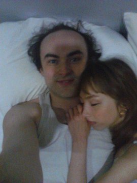 A bearded man reclines against a white pillow, gazing softly at the camera while a young woman with brown hair rests her head on his shoulder with her eyes closed. The intimate, dimly lit photo appears to be a personal moment, though it is unclear how it relates to the accompanying post about William Banks and the described events.
