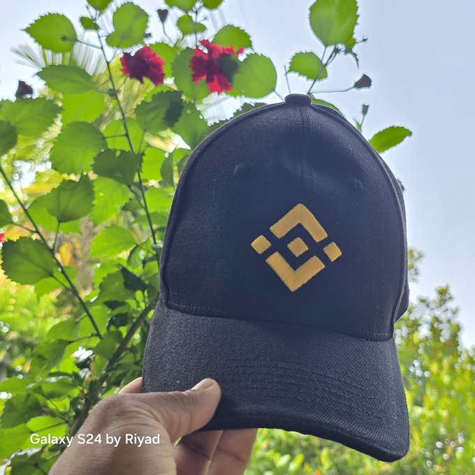 The image features a person holding up a black baseball cap with a yellow diamond design, which has the word "Binance" written on it. The caption of the image reads "Good Morning, X Family Starting the day with my favorite Binance cap and a positive mindset. Wishing everyone a productive day filled with opportunities, growth, and success."