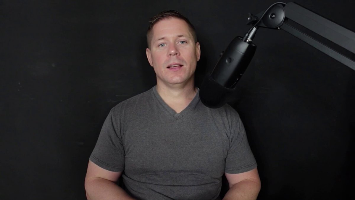 The image shows a man wearing a gray shirt sitting behind a microphone with his hands resting on his knees. The background of the image is black, which contrasts with the man's attire. The focus of the image seems to be on the man rather than the surrounding environment or any other objects in the scene.