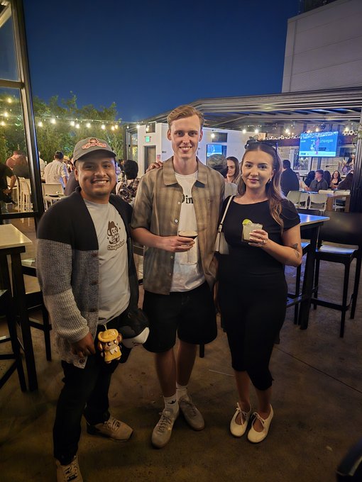 Three friends posing together at an outdoor evening venue, with one holding a drink and a small plush toy.