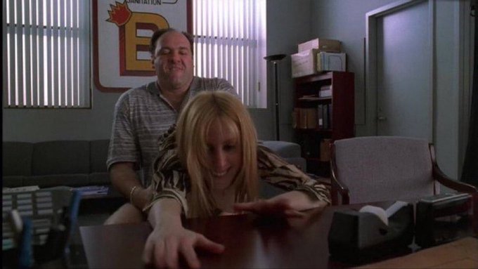 A man stands behind a blonde woman leaning over a desk in an office, with a "MANDATION" poster visible on the wall behind them. The image humorously illustrates the post's message about the crypto industry's collapse, depicting a physically intimate yet absurd scenario where the woman appears to be in a compromising position—mirroring the "dead and never coming back" sentiment about crypto investments through this awkward, unflattering visual metaphor.