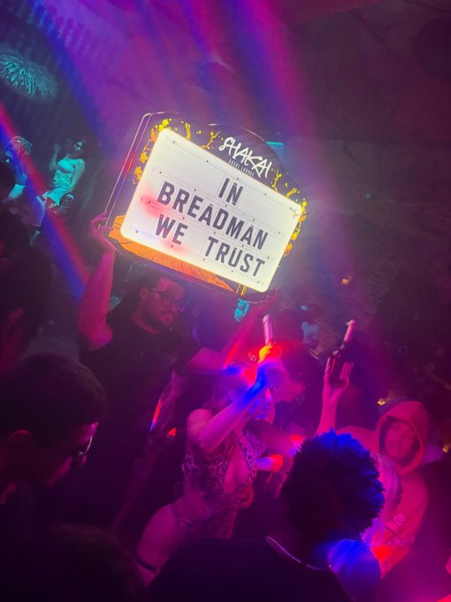 A nighttime club or party scene where someone holds up an illuminated sign reading 'IN BREADMAN WE TRUST' with 'SHAKAI CLUB IBARAKI' branding, as the crowd dances under pink and purple lighting.