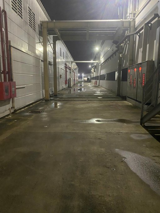 A wet industrial alleyway runs between buildings at night, illuminated by overhead lights reflecting off the puddles on the concrete floor.