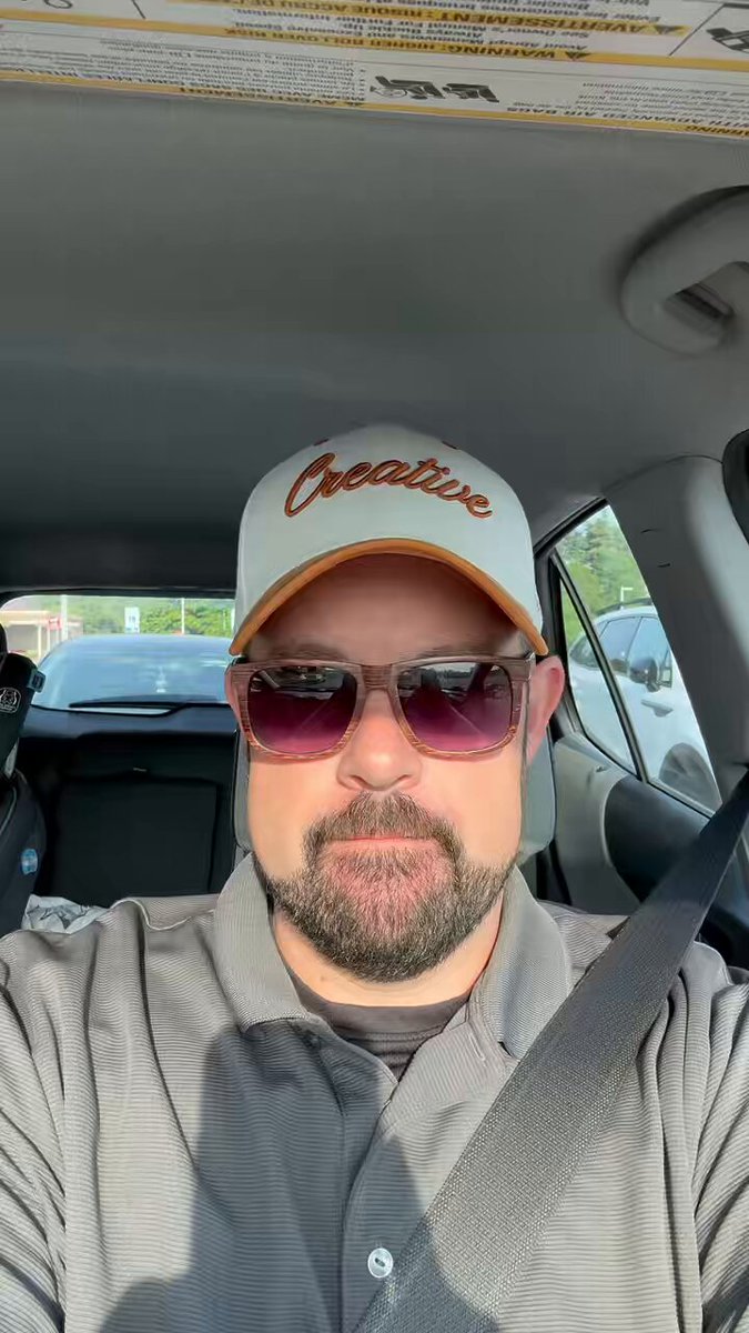 The image features a man wearing sunglasses, a gray shirt, and a white baseball cap with "Creative" written on it. He appears to be sitting inside a car, possibly driving or waiting at a stop sign. The background of the photo shows several cars parked in various positions, indicating that he is likely in an urban area where parking spots are limited. There's also a post about a store called Target located near his current location, which might have some relevance to him if he's on his way there or just arrived from one.