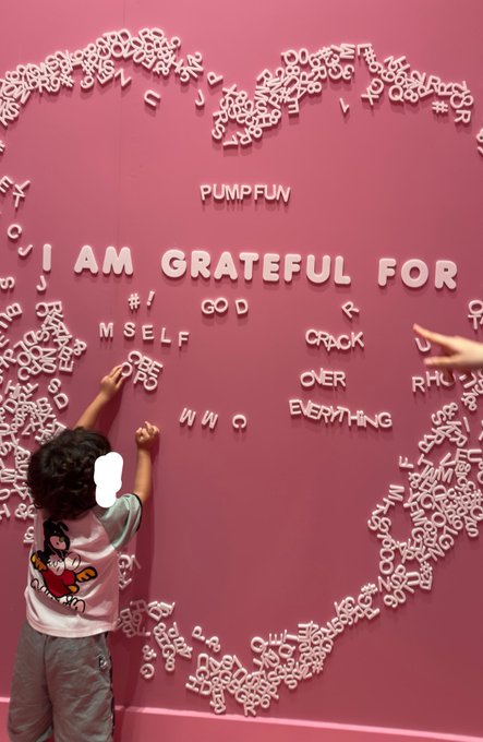 A child in a Mario shirt places white letters on a pink "I AM GRATEFUL FOR" heart-shaped wall, arranging words like "PUMP FUN" and "MYSELF" to create a heartfelt message of gratitude. The installation visually echoes the post's sentiment of thankfulness, with the "PUMP FUN" ticker prominently displayed among the crafted sentiments.