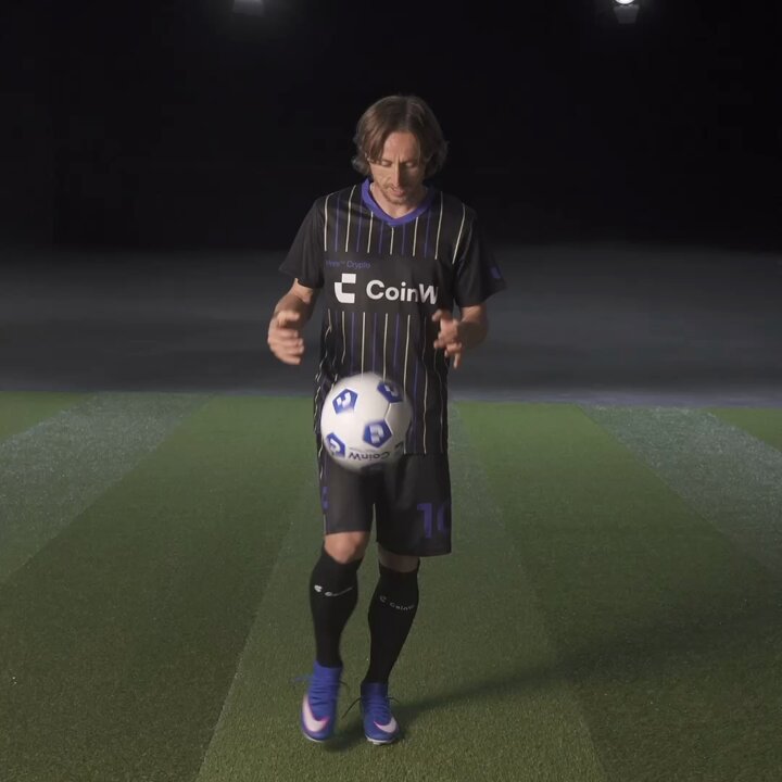 The image features a man dressed in soccer attire, holding a soccer ball and preparing to juggle it. The background of the photo shows a dark room with a green field visible through a window or door. There are also several other people present in the scene, but they do not appear to be involved in the juggling activity.