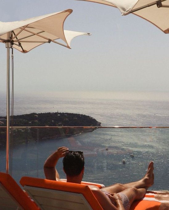 The image shows a man sitting on an orange lounge chair, looking out at the ocean from his balcony. The man appears relaxed as he enjoys the view of the water below. In the background, there are several boats visible near the shoreline. The scene is set against a clear blue sky and calm waters, creating a serene atmosphere for the man to unwind in his lounge chair.