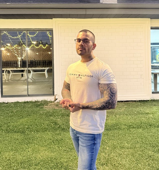 A man with tattoos, wearing a white Tommy Hilfiger t-shirt and blue jeans, stands outside a house with white brick walls and string lights visible through the window.