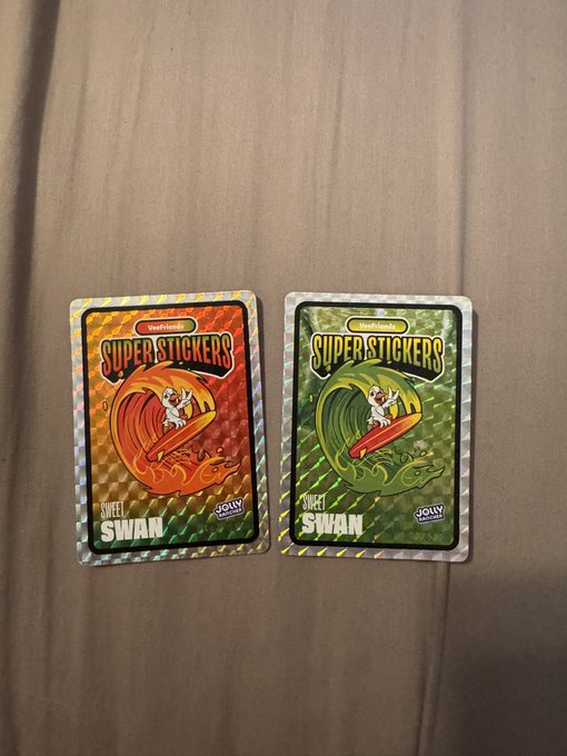 Two Hearthstone 'Superstickers' collectible card packs from the 'Sweet Swan' series lie on a tan fabric surface, one with an orange flame design and the other with a green dragon design, both made by Joki.
