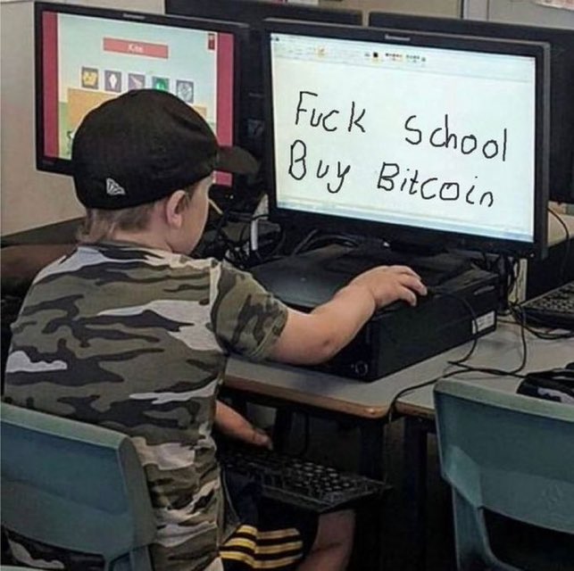 A young child wearing a backward cap and camouflage shirt sits at a computer displaying the handwritten message 'Fuck School Buy Bitcoin' on the monitor.