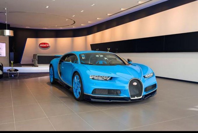 A blue Bugatti Chiron is displayed on the showroom floor of a luxury car dealership, with the Bugatti logo visible on the wall in the background, representing the kind of extravagant purchase the user jokingly considered after a massive crypto trading win.