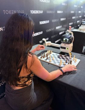 A woman plays chess against a humanoid robot at an event branded with "TOKEN2049" logos on the backdrop.