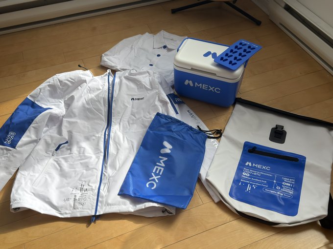A collection of MEXC-branded merchandise is laid out on a wooden floor, including a white windbreaker jacket, a polo shirt, a cooler bag, and a backpack.