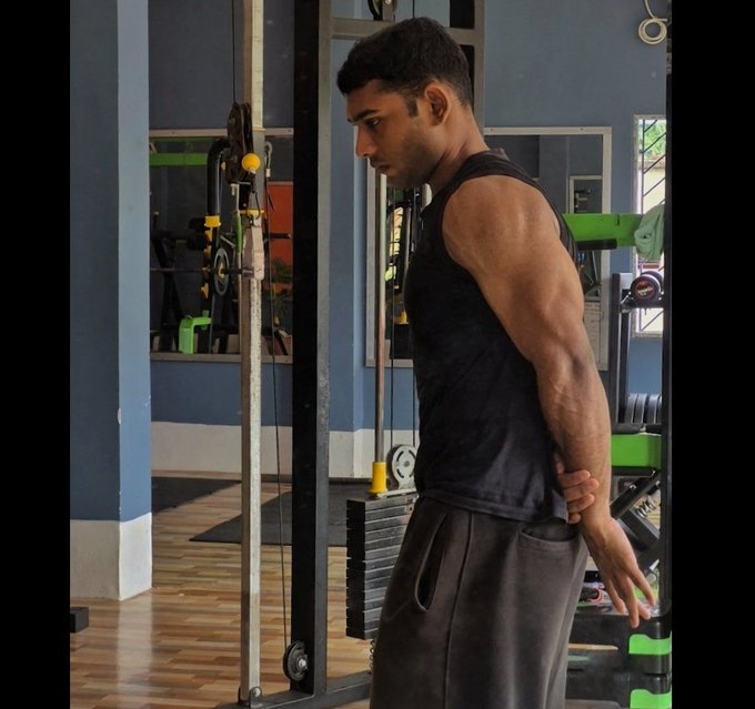 The image shows a man wearing a black tank top, gray sweatpants, and holding his arm out while standing next to a weight machine at a gym. The surrounding area appears to be well-equipped with various exercise equipment, including dumbbells and a bench press. The focus of the post is on the man's arms, suggesting that he might be working on building or maintaining muscle mass.