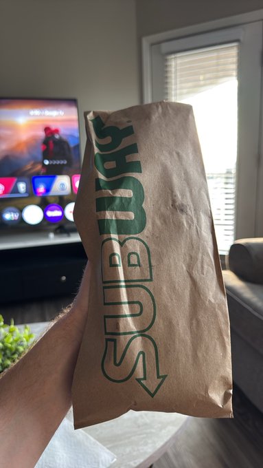 A first-person view of someone holding a brown paper bag with green "SUBWAY" branding, with an Apple TV screen visible in the background.