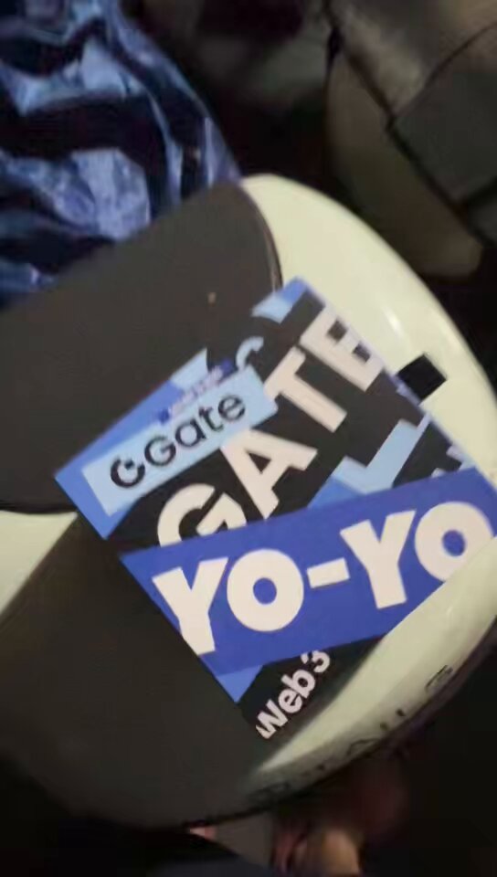 The image features a white chair with a blue sticker that says "gate" on it, along with the words "yo-yo", "gatechild", and "cex". The chair appears to be made of wood or plastic material. There is also a person's hand visible in the bottom right corner of the photo.