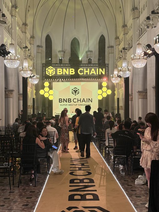 The "BNB CHAIN Super Meetup Singapore" event attracted a large crowd inside an ornate hall, with attendees seated along a golden runway displaying "BNB CHAIN" branding beneath the event's illuminated backdrop, reflecting the post's observation about the consistently large turnouts.