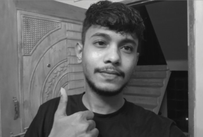A black-and-white selfie video frame of a young man giving a thumbs-up gesture in front of an ornate door and staircase.